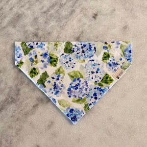 May include: A triangular dog bandana featuring a watercolor floral pattern of blue and purple hydrangeas with green leaves on a white background. The bandana has a small tag on the right side.