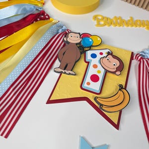 May include: A colorful birthday banner featuring a cartoon monkey holding balloons, a number "1", and a banana. The banner has red, white, yellow, and blue ribbons. The word "Birthday" is also visible.
