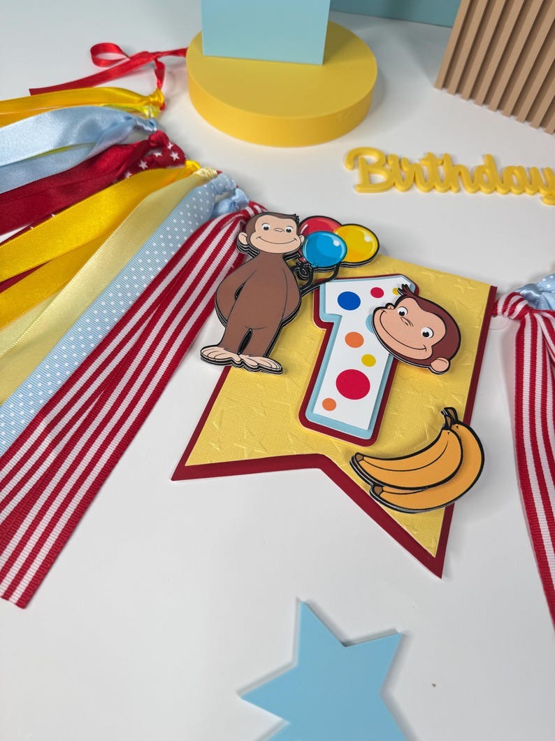 May include: A colorful birthday banner featuring a cartoon monkey holding balloons, a number "1", and a banana. The banner has red, white, yellow, and blue ribbons. The word "Birthday" is also visible.