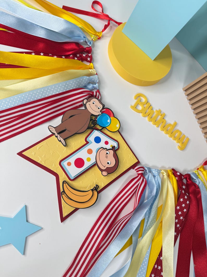 May include: A colorful birthday banner with ribbons in red, yellow, and blue. The banner features a cartoon monkey, a number one, and the word "Birthday". Decorative elements include a star and a banana.