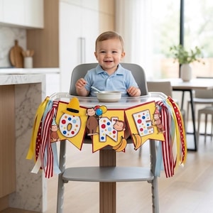 May include: A smiling baby sits in a high chair with a colorful banner that says "ONE". The banner features cartoon monkeys, bananas, and polka dots. Ribbons in yellow, red, and blue decorate the high chair.