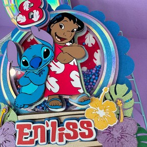Może przedstawiać: Kolorowa, warstwowa dekoracja na tort z postaciami Lilo i Stitch. Projekt zawiera cyfrę 8, imię "Enliss" i tropikalne kwiaty. Ozdoba jest ozdobiona brokatem i r&oacute;żnymi odcieniami niebieskiego, czerwonego i ż&oacute;łtego.