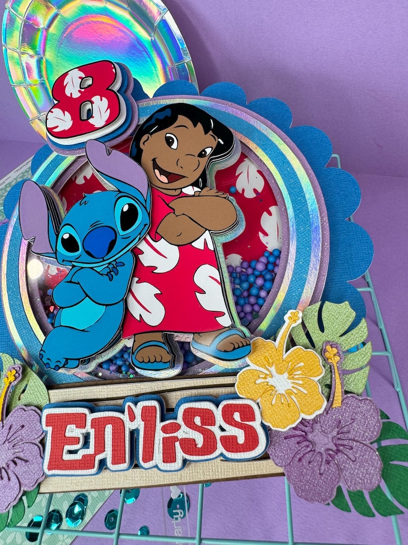 Może przedstawiać: Kolorowa, warstwowa dekoracja na tort z postaciami Lilo i Stitch. Projekt zawiera cyfrę 8, imię "Enliss" i tropikalne kwiaty. Ozdoba jest ozdobiona brokatem i r&oacute;żnymi odcieniami niebieskiego, czerwonego i ż&oacute;łtego.