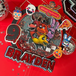 May include: A red and black cake topper with the name "BRAYDEN" in red letters. The topper features characters from the video game "Five Nights at Freddy's", including Freddy Fazbear, Foxy, and Bonnie. The number "9" is also included.