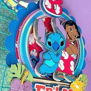 Może przedstawiać: Kolorowa, warstwowa dekoracja na tort z postaciami Stitcha i Lilo. Projekt zawiera cyfrę osiem, tęczę i napis "Enliss". Ozdobne kwiaty i liście dopełniają świąteczny motyw.