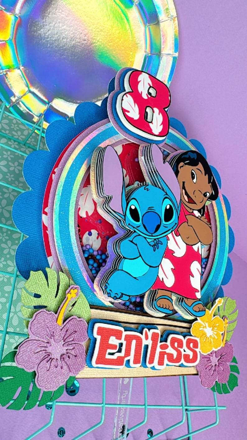 Może przedstawiać: Kolorowa, warstwowa dekoracja na tort z postaciami Stitcha i Lilo. Projekt zawiera cyfrę osiem, tęczę i napis "Enliss". Ozdobne kwiaty i liście dopełniają świąteczny motyw.