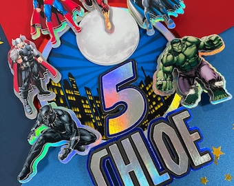 May include: A cake topper featuring a blue and white cityscape with a moon, the number 5, and the name CHLOE in iridescent letters.  The topper also includes superhero figures: Superman, Batman, The Flash, Thor, Hulk, and Black Panther.