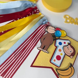 May include: A festive banner with red, yellow, and blue ribbons. A banner with a cartoon monkey holding balloons, a number 1, and a banana. The banner is yellow with a red border. Birthday decorations.