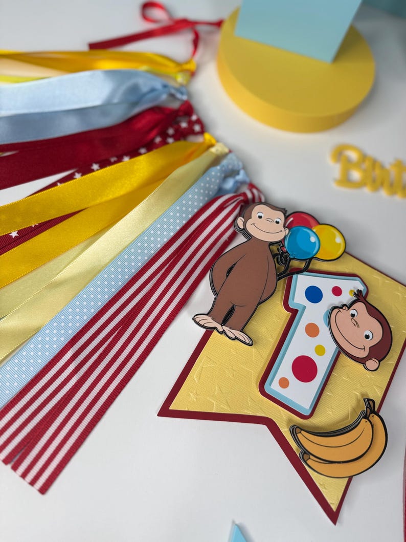 May include: A festive banner with red, yellow, and blue ribbons. A banner with a cartoon monkey holding balloons, a number 1, and a banana. The banner is yellow with a red border. Birthday decorations.