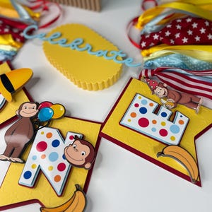 May include: A festive birthday banner featuring a yellow background with red trim, adorned with the letter "N" and a cartoon monkey. The banner includes a banana and colorful polka dots. A yellow cake-shaped decoration with the word "celebrate" is also visible.