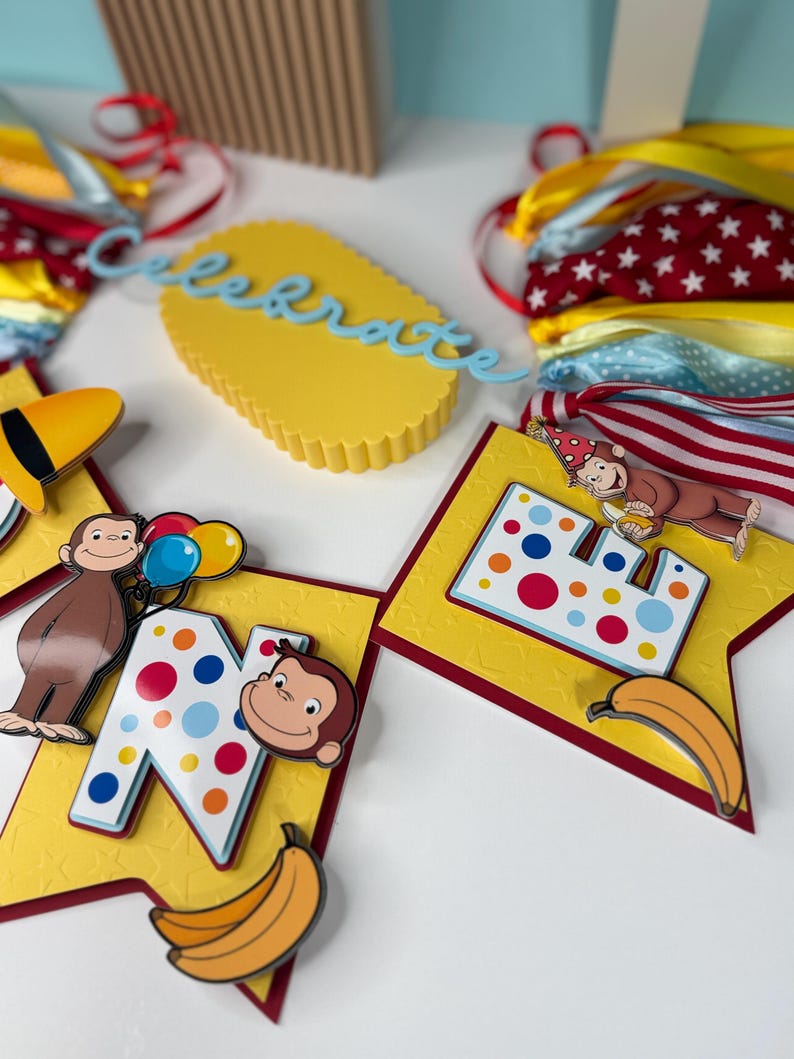 May include: A festive birthday banner featuring a yellow background with red trim, adorned with the letter "N" and a cartoon monkey. The banner includes a banana and colorful polka dots. A yellow cake-shaped decoration with the word "celebrate" is also visible.