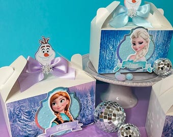 May include: White gable boxes with Frozen-themed designs. Each box features a character image, a ribbon, and an Olaf topper. The boxes are decorated with images of Elsa and Anna, and are accompanied by disco balls and pom-poms.