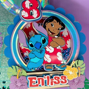 Może przedstawiać: Kolorowa dekoracja na tort z postaciami Stitcha i Lilo. Numer 8 jest wyświetlany w kolorze czerwono-białym, poniżej znajduje się napis "Eniiss". Ozdoba jest udekorowana kwiatami i liśćmi.