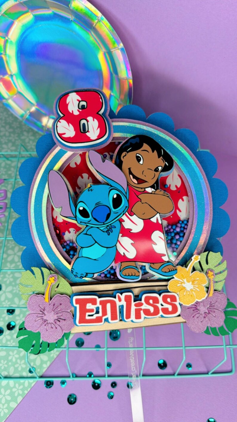 Może przedstawiać: Kolorowa dekoracja na tort z postaciami Stitcha i Lilo. Numer 8 jest wyświetlany w kolorze czerwono-białym, poniżej znajduje się napis "Eniiss". Ozdoba jest udekorowana kwiatami i liśćmi.