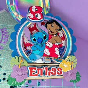 Może przedstawiać: Kolorowa dekoracja na tort urodzinowy z postaciami Stitcha i Lilo z filmu Disneya Lilo & Stitch. Ozdoba zawiera cyfrę 8, imię "Enilss" i dekoracyjne kwiaty. Tło to mieszanka fioletowych, niebieskich i iryzujących element&oacute;w.