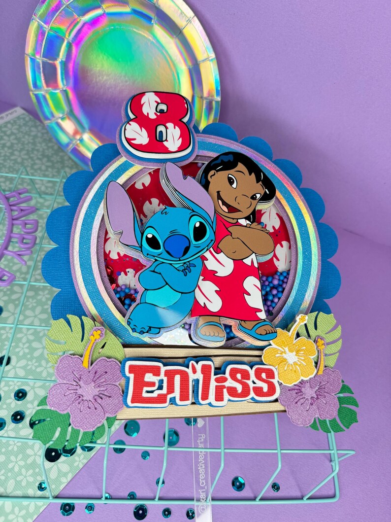 Może przedstawiać: Kolorowa dekoracja na tort urodzinowy z postaciami Stitcha i Lilo z filmu Disneya Lilo & Stitch. Ozdoba zawiera cyfrę 8, imię "Enilss" i dekoracyjne kwiaty. Tło to mieszanka fioletowych, niebieskich i iryzujących element&oacute;w.