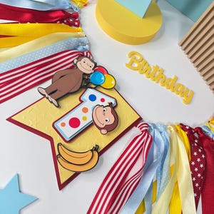 May include: A colorful birthday banner featuring a cartoon monkey holding balloons, a number one, and bananas. The banner includes red, yellow, and blue ribbons, and the word "Birthday".