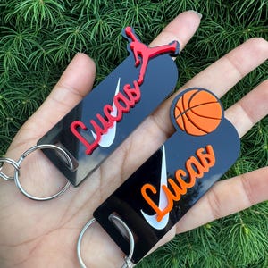 May include: Two black keychains with white Nike swoosh logos and the name "Lucas" in orange and red lettering. One keychain features a red basketball player silhouette and the other features an orange basketball.