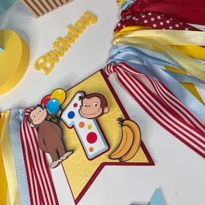 May include: A festive birthday banner with colorful ribbons in red, yellow, and blue. The banner features a number one, a cartoon monkey holding balloons, and bananas. The word "Birthday" is also visible.