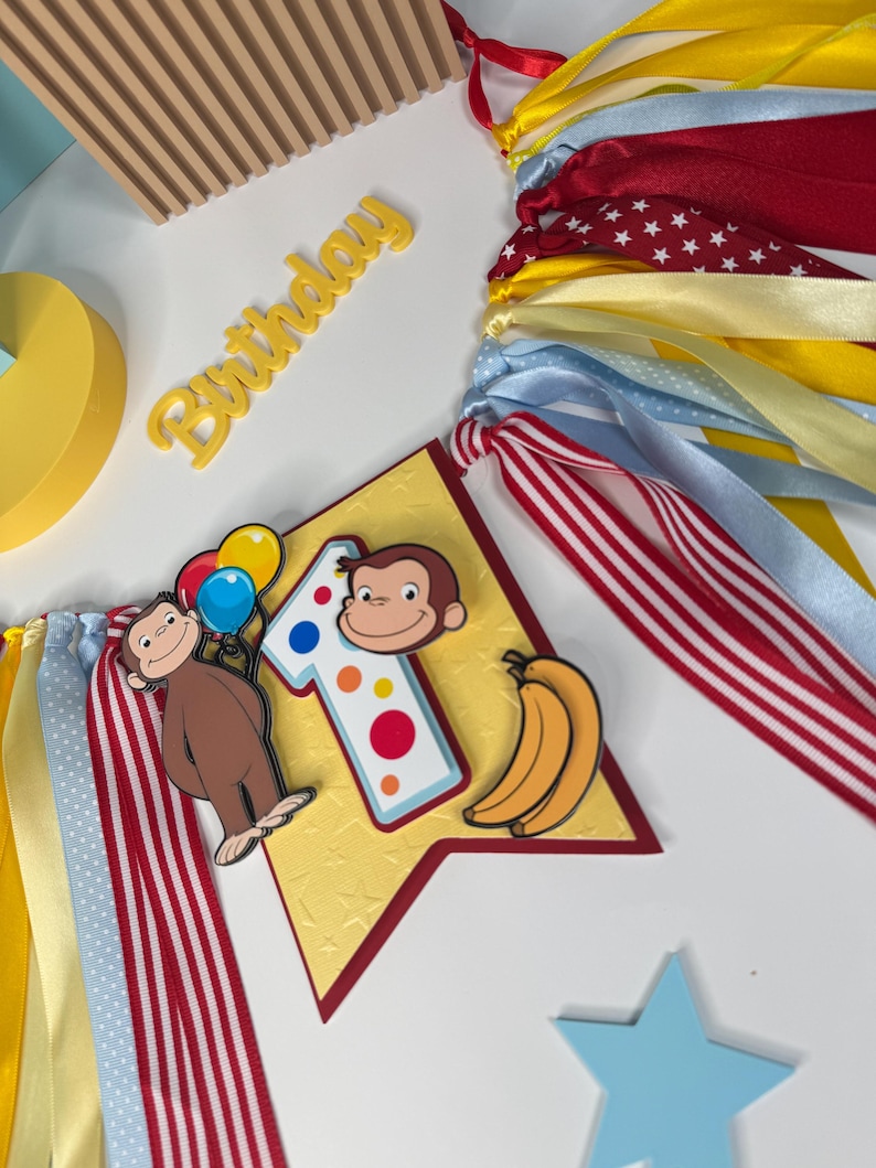 May include: A festive birthday banner with colorful ribbons in red, yellow, and blue. The banner features a number one, a cartoon monkey holding balloons, and bananas. The word "Birthday" is also visible.