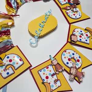May include: A yellow banner with red trim and the word "CELEBRATE" in blue script. The banner features the letters "E", "O", and "B" with colorful polka dots and cartoon monkey illustrations. Ribbons in various colors are attached.