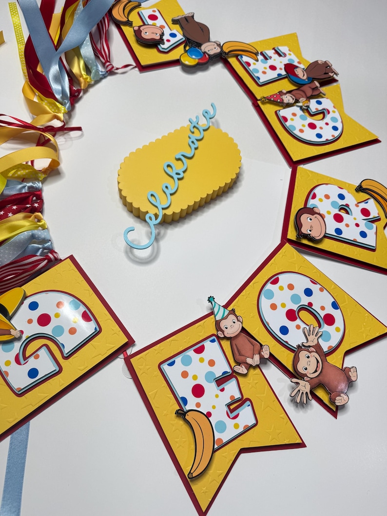 May include: A yellow banner with red trim and the word "CELEBRATE" in blue script. The banner features the letters "E", "O", and "B" with colorful polka dots and cartoon monkey illustrations. Ribbons in various colors are attached.