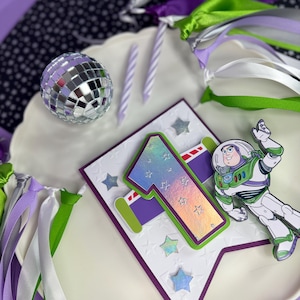 May include: A festive birthday scene with a disco ball, candles, and a banner featuring the number "1" and a Buzz Lightyear figure. The banner is white with embossed stars and a purple border. Ribbons in green, purple, and silver add to the celebratory feel.