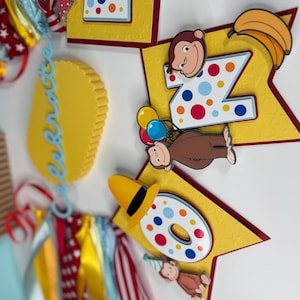 May include: A colorful birthday banner featuring the number "2" and "0" with a cartoon monkey theme. The banner includes yellow star-shaped cutouts, bananas, and the word "Celebrate" in blue script. Ribbons in red, white, and yellow add to the festive design.