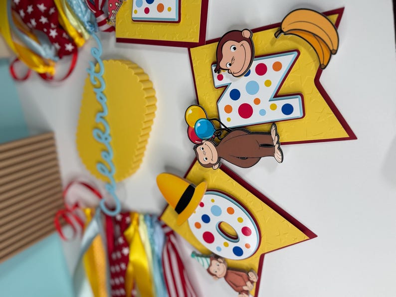 May include: A colorful birthday banner featuring the number "2" and "0" with a cartoon monkey theme. The banner includes yellow star-shaped cutouts, bananas, and the word "Celebrate" in blue script. Ribbons in red, white, and yellow add to the festive design.