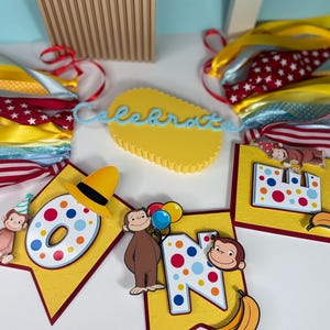 May include: A festive banner with the word "ONE" and a monkey theme. The banner features yellow pennants with red borders, colorful polka dots, and illustrations of a monkey holding balloons and bananas. A yellow cake topper with the word "Celebrate" is also visible.