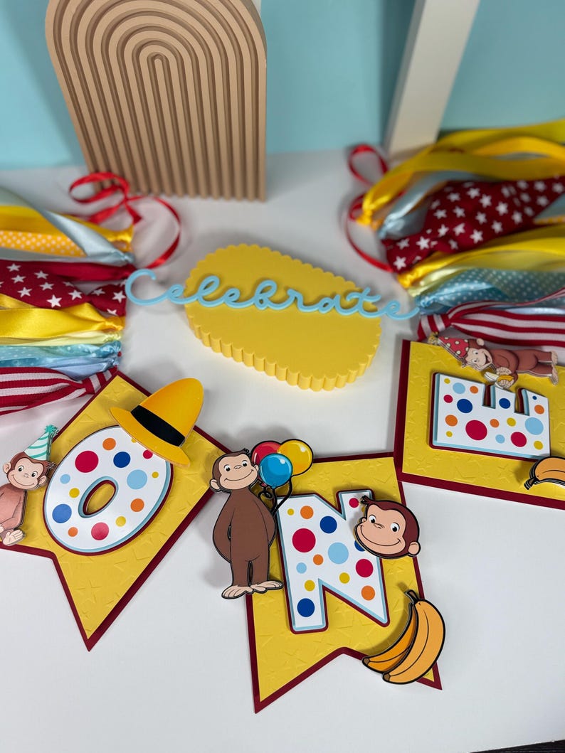 May include: A festive banner with the word "ONE" and a monkey theme. The banner features yellow pennants with red borders, colorful polka dots, and illustrations of a monkey holding balloons and bananas. A yellow cake topper with the word "Celebrate" is also visible.