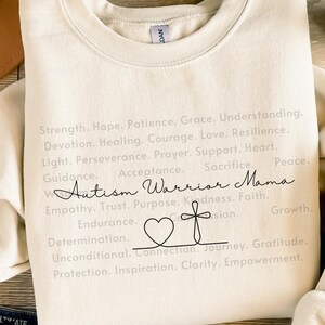 May include: A cream-coloured sweatshirt with the words "Autism Warrior Mama" in a handwritten font, accompanied by a heart and cross graphic. The sweatshirt also has a list of words such as "Strength," "Hope," and "Love."