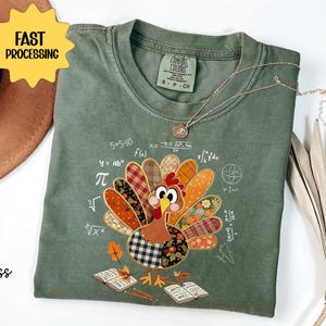 May include: A moss green t-shirt with a colourful turkey design featuring a patchwork pattern. The turkey is surrounded by mathematical symbols and equations. A gold necklace is draped on the shirt.
