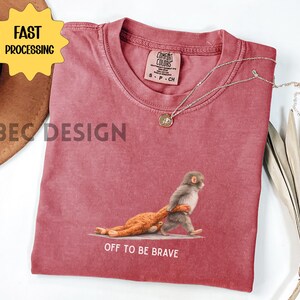 May include: A red Comfort Colors t-shirt featuring a graphic of a monkey carrying a stuffed animal, with the text "OFF TO BE BRAVE". A gold necklace and a brown hat are also visible. The shirt has a vintage look.