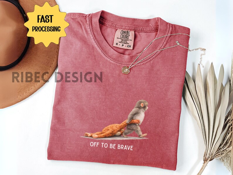 May include: A red Comfort Colors t-shirt featuring a graphic of a monkey carrying a stuffed animal, with the text "OFF TO BE BRAVE". A gold necklace and a brown hat are also visible. The shirt has a vintage look.