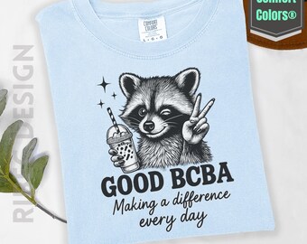 May include: Light blue t-shirt with a raccoon graphic holding a boba drink and making a peace sign. The text reads "GOOD BCBA Making a difference every day." The shirt is labeled "Comfort Colors".
