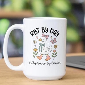 May include: White ceramic mug featuring a cartoon goose with a pink bow, boots, and the text "RBT BY DAY" and "Silly Goose by Choice." The mug has a curved handle and is decorated with flowers and a butterfly.