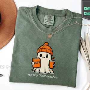 May include: A moss green Comfort Colors t-shirt with a cartoon ghost wearing an orange beanie, holding a math book and a drink. The text "Spooky Math Teacher" is below the graphic. A gold necklace is draped on the shirt.