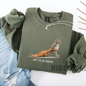 May include: An olive green sweatshirt with a graphic of a monkey carrying a stuffed animal, with the text "OFF TO BE BRAVE". A silver necklace is draped across the sweatshirt. The sweatshirt is paired with blue jeans.