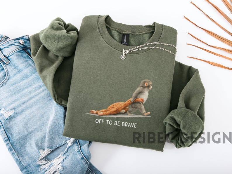 May include: An olive green sweatshirt with a graphic of a monkey carrying a stuffed animal, with the text "OFF TO BE BRAVE". A silver necklace is draped across the sweatshirt. The sweatshirt is paired with blue jeans.