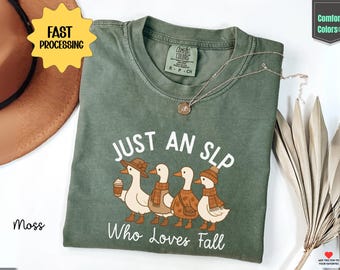 May include: A moss green Comfort Colors t-shirt with the text "JUST AN SLP Who Loves Fall" and a graphic of four ducks wearing fall attire. A gold necklace is draped on the shirt.