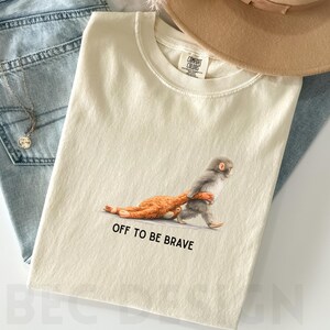 May include: Ivory-colored t-shirt featuring a whimsical illustration of a monkey carrying a smaller animal, with the text "OFF TO BE BRAVE" printed below. The shirt is paired with blue jeans and a tan hat.