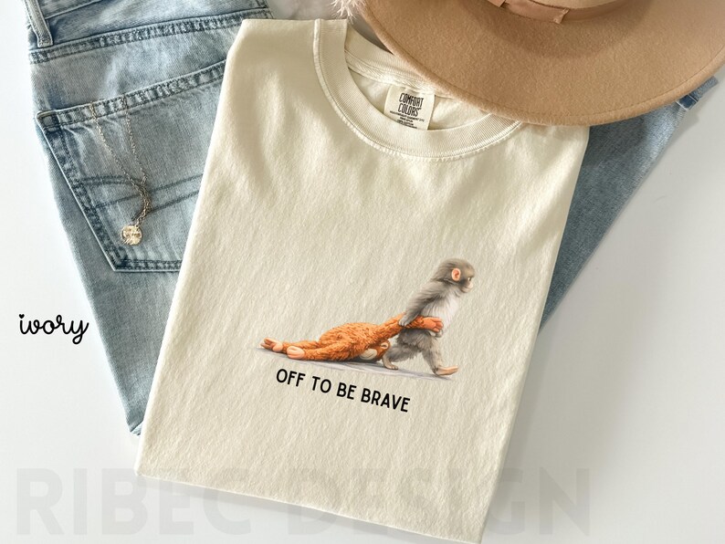 May include: Ivory-colored t-shirt featuring a whimsical illustration of a monkey carrying a smaller animal, with the text "OFF TO BE BRAVE" printed below. The shirt is paired with blue jeans and a tan hat.