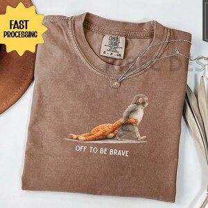 May include: Brown t-shirt with a graphic of a monkey carrying a stuffed animal, with the text "OFF TO BE BRAVE". The shirt has a crew neck and short sleeves. A silver necklace with a pendant is on the shirt.