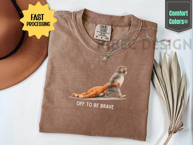 May include: Brown t-shirt with a graphic of a monkey carrying a stuffed animal, with the text "OFF TO BE BRAVE". The shirt has a crew neck and short sleeves. A silver necklace with a pendant is on the shirt.