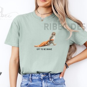 May include: A light green t-shirt with a graphic of a monkey carrying a chicken, with the text "OFF TO BE BRAVE". The shirt is made of a soft material.