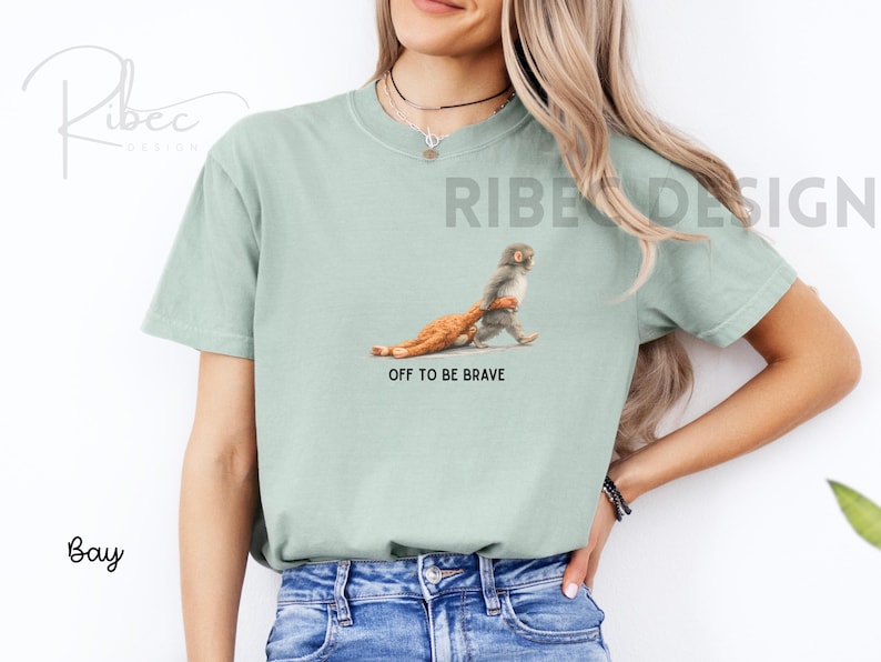 May include: A light green t-shirt with a graphic of a monkey carrying a chicken, with the text "OFF TO BE BRAVE". The shirt is made of a soft material.