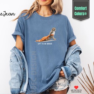 May include: Blue jean-colored t-shirt with the text "OFF TO BE BRAVE" and a graphic of a monkey and a lion. The shirt is part of the Comfort Colors collection.
