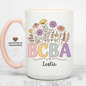 Könnte beinhalten: Weißer Keramikbecher mit rosa Griff und Innenraum. Der Becher zeigt die Buchstaben "BCBA" in Pastellfarben, Blumenillustrationen und den Namen "Leslie" in schwarzer Schrift. Auf dem Griff befindet sich ein Herzsymbol.