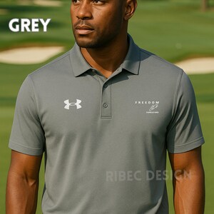 Op de afbeelding: Een grijze poloshirt met een wit logo op de linkerborst en het woord "FREEDOM" op de rechterborst. De shirt heeft een knoopsluiting met drie knopen en korte mouwen. Het woord "GREY" staat bovenaan de afbeelding.